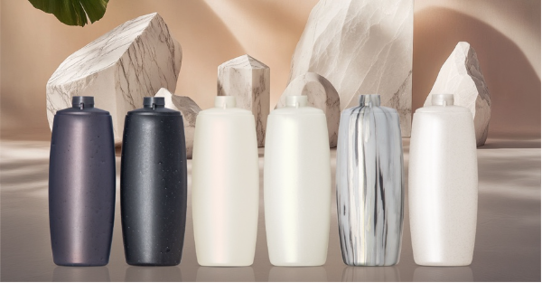 Silken Stone | AMPACET CORPORATION Row of six tall cosmetic bottles in varying colors purple dark blue white white marbled white lined up on a reflective surface with a rocky beige backdrop
